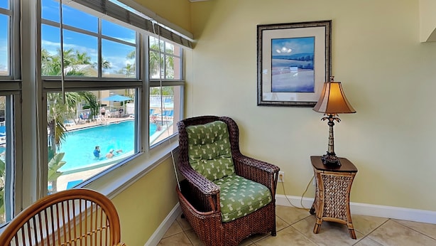 Una habitación con vista a una piscina, una silla de mimbre, un cuadro en la pared y una lámpara de mesa.