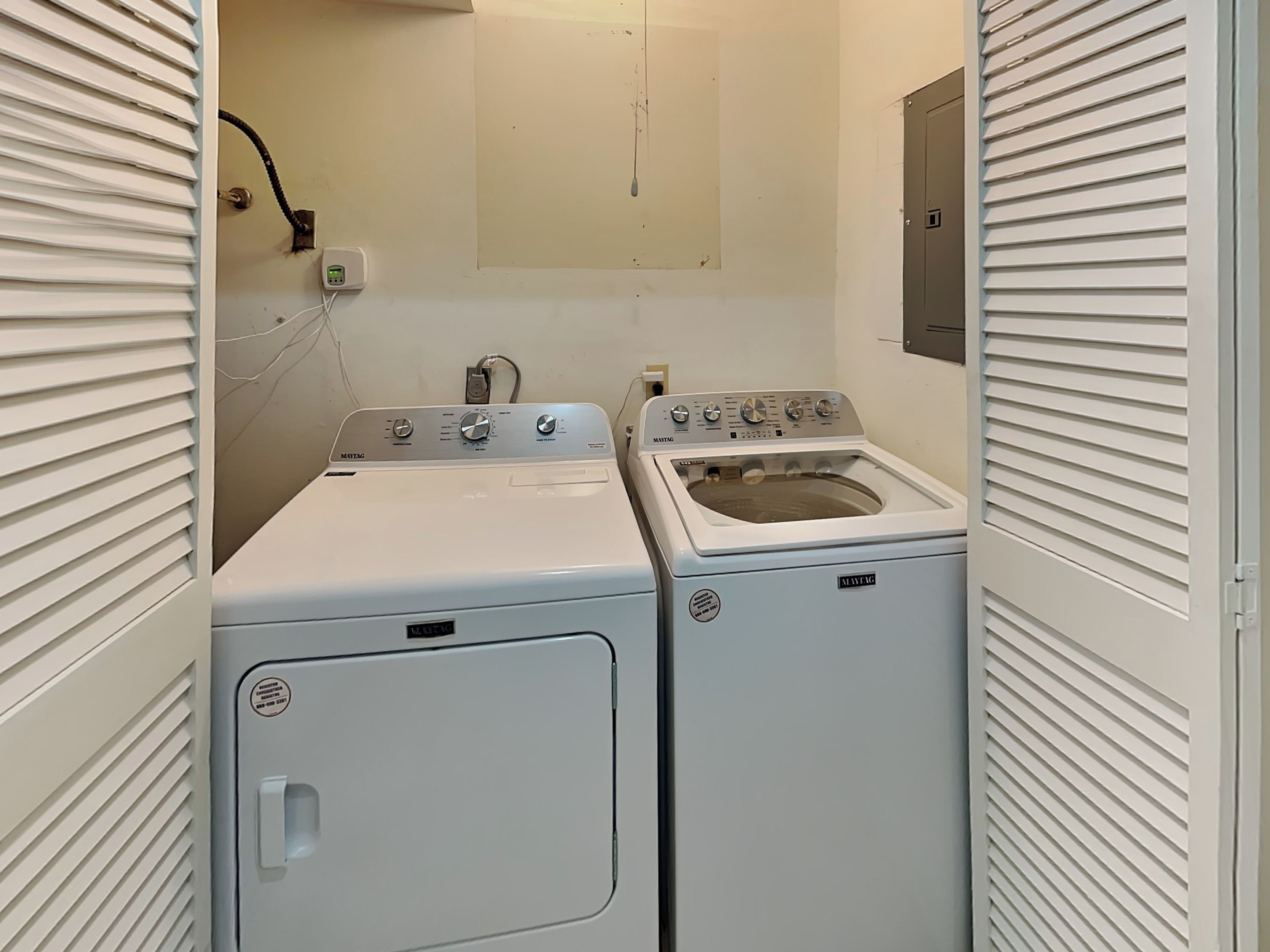 A laundry room with two white front-loading washers and dryers, a control panel, and a wall-mounted electrical outlet.