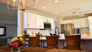 A modern kitchen with white cabinets, stainless steel appliances, and a dining area with wicker chairs and a vase of flowers. - Margarita House in The Villages close to both Lake Sumpter and Spanish Springs! (The Villages)
