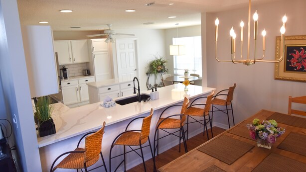 A modern kitchen with white cabinets, a central island with bar stools, and a dining area with a wooden table and woven chairs. - Margarita House in The Villages close to both Lake Sumpter and Spanish Springs! (The Villages)