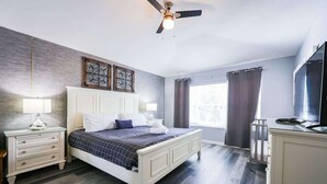 Ein Schlafzimmer mit Bett, Nachttischen, Deckenventilator, einem Fenster mit Vorhängen und einem Fernseher.