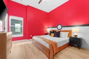 Un dormitorio con pared roja, una cama de madera, una mesita de noche con lámpara y una ventana con persianas.