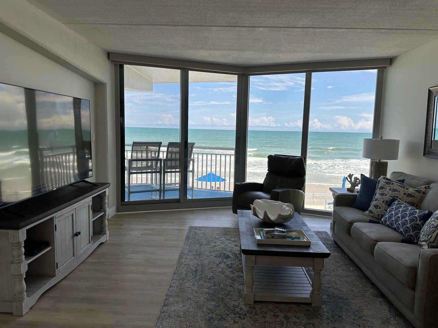 A living room with a large TV, a sofa, a coffee table, and a view of the ocean through large windows.