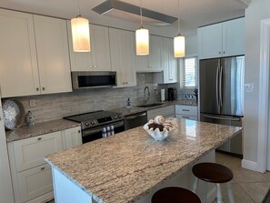 Una cocina moderna con armarios blancos, encimera de granito, electrodomésticos de acero inoxidable e iluminación colgante.