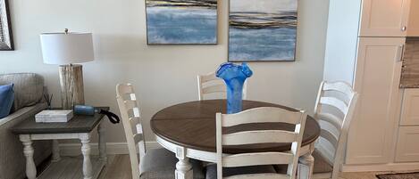 Un comedor con una mesa redonda, sillas blancas, un jarrón azul y dos cuadros abstractos en la pared.