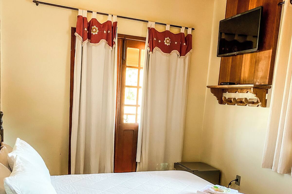 A bedroom with a bed, curtains, a television mounted on the wall, and a wooden ceiling.