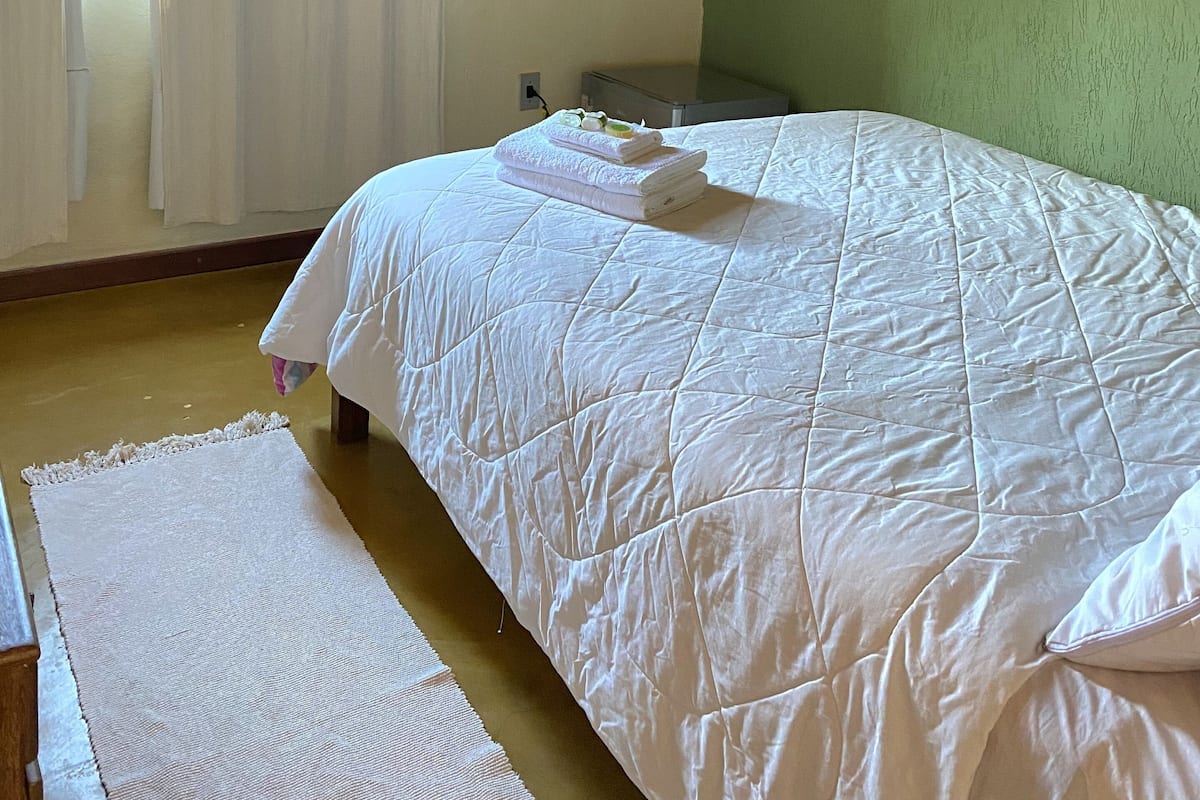 A neatly made bed with a quilted white duvet, a wooden headboard, a flat-screen TV mounted above it, and a window with white curtains.