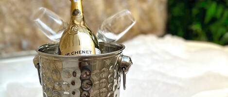 Una botella de champán en un cubo metálico decorativo con asas, sobre una superficie blanca con un muro de piedra y follaje al fondo.