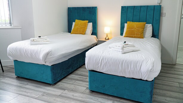 Two twin beds with teal headboards and white bedding, a bedside table with a lamp, and a window with blinds. - Central twin bed flat (Cardiff)