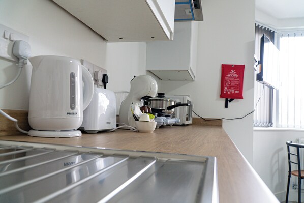 A kitchen with a Prolex coffee maker, a kettle, and a toaster. There is a fire safety sign on the wall and a window with blinds.