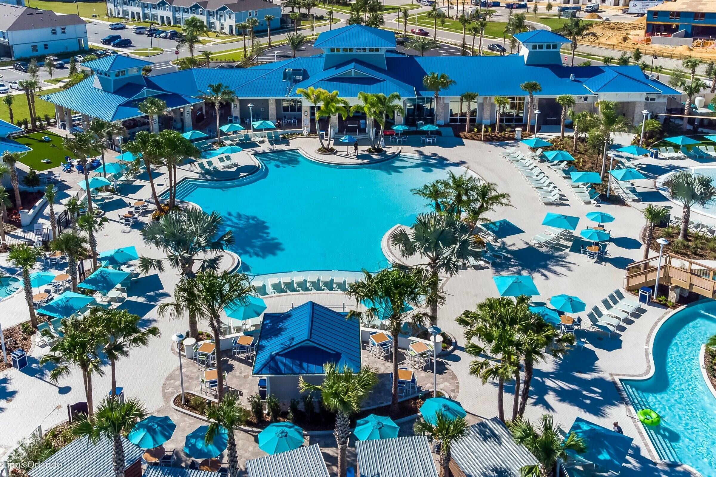 An aerial view of a resort with a large central pool, multiple lounge areas with umbrellas, and palm trees.