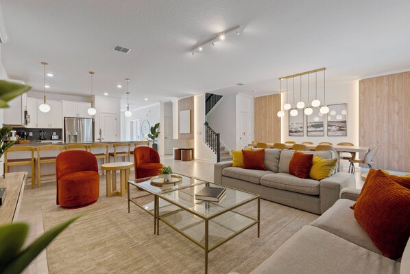 A modern living room with a sofa, coffee table, and chairs. There is a kitchen area with cabinets and a dining table.
