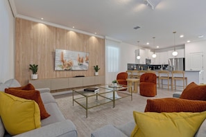 Ein modernes Wohnzimmer mit einer Couch, Sesseln und einem Couchtisch. Im Hintergrund befindet sich eine Küchenzeile mit Schränken und Geräten.