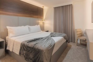 Una habitación de hotel moderna con una cama grande, una mesita de noche, una silla y un ventanal con cortinas.