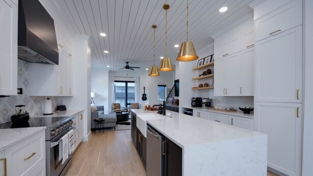 Een moderne keuken met witte kasten, een groot eiland, hanglampen en een zichtbare trap die naar een andere verdieping leidt.