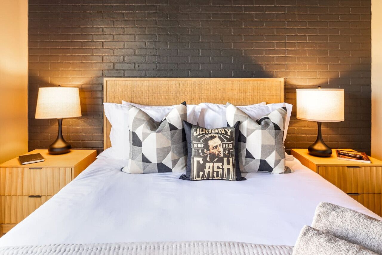 Une chambre d’hôtel avec un mur en briques, une tête de lit en bois, deux tables de chevet avec des lampes et un lit avec une literie blanche et des oreillers à motifs.