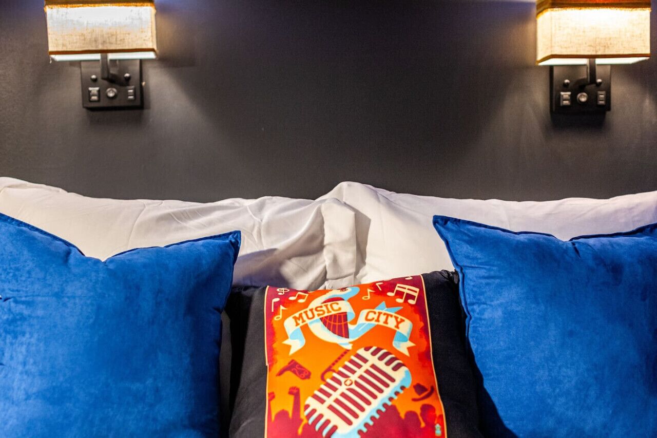 Un lit avec des draps blancs, des oreillers bleus et un coussin orné d’un motif représentant la ville de la musique.