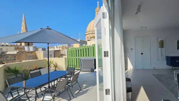 En balkon med et bord, stole, en sofa og udsigt over bybilledet, inklusive et kirketårn.