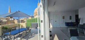 A balcony with a table and chairs, a sofa, and a view of a cityscape including a church tower. - Valletta Penthouse Cathedral view Terrace (Valletta)