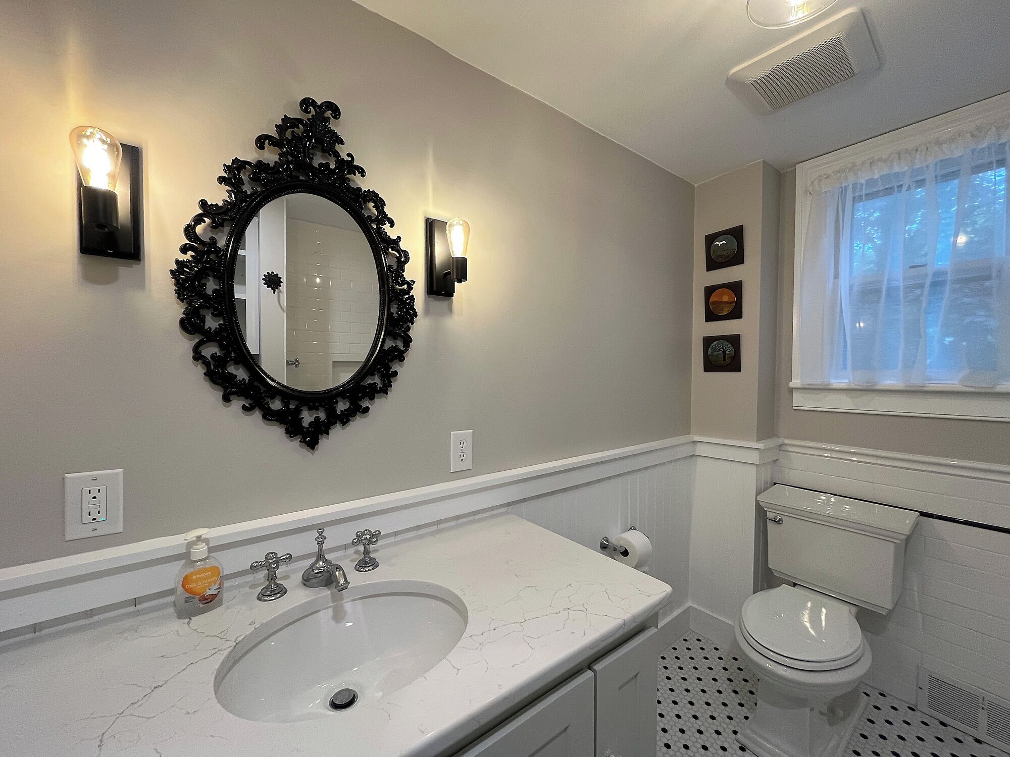 A bathroom with a marble sink, a black ornate mirror, wall sconces, a toilet, and a window with sheer curtains.