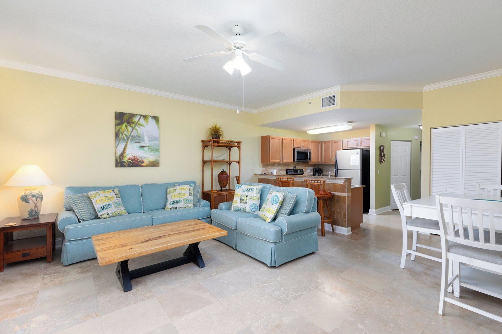 A living room with a sofa set, a coffee table, a dining table with chairs, a kitchen area with cabinets and appliances, and a ceiling fan.