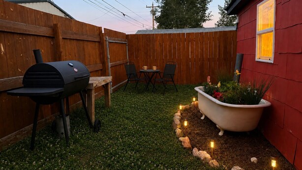 A backyard with a barbecue grill, a small table with chairs, a potted plant, and a red building.