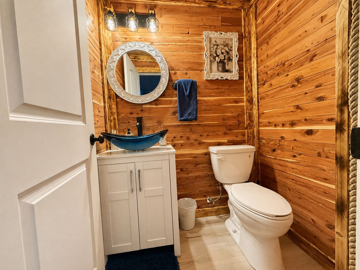 Un baño con paredes de madera, un inodoro blanco, un lavamanos azul con un cuenco decorativo, un espejo con un marco ornamentado y un pequeño cubo de basura.
