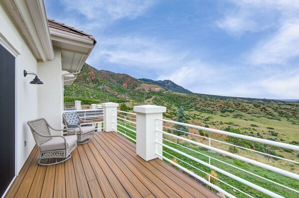 A wooden deck with outdoor furniture, a white railing, and a scenic view of green hills and mountains.