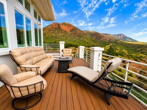 A deck with outdoor furniture, a view of a mountainous landscape, and a building with large windows. - Luxury Mountain-View Retreat | near Garden of the Gods | Hot Tub | Game Room (Colorado Springs)