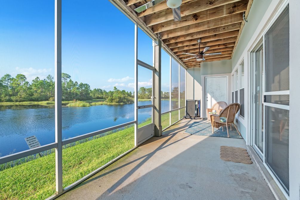 A covered balcony with large windows overlooking a lake, a ceiling fan, and a wicker chair.
