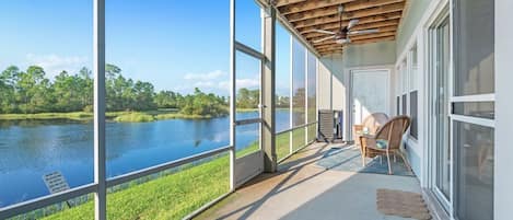 Ein überdachter Balkon mit großen Fenstern und Blick auf einen See, ein Deckenventilator und ein Korbstuhl.