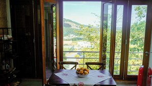 Un coin repas avec une table dressée pour deux personnes, un saladier rempli de fruits et une vue sur un paysage verdoyant, visible à travers de grandes portes vitrées.