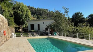 Une piscine rectangulaire à l’eau claire, entourée d’une terrasse carrelée, avec un bâtiment blanc en arrière-plan et des arbres.