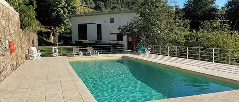 Une piscine rectangulaire à l’eau claire, entourée d’une terrasse carrelée, avec un bâtiment blanc en arrière-plan et des arbres.