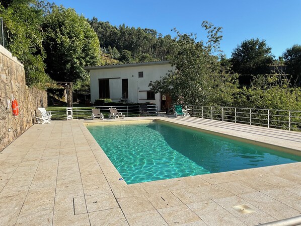 A rectangular pool with clear water, surrounded by a tiled deck, a white building in the background, and trees.