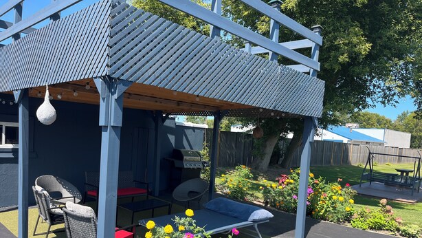 A covered outdoor seating area with a grill, wicker chairs, and a potted plant with flowers. - East Court Motel (Sarnia)