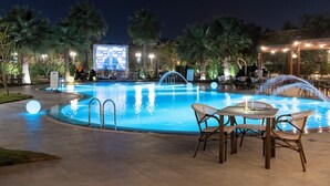Une piscine entourée d’un patio comprenant une table et des chaises, des palmiers et un grand écran lumineux.