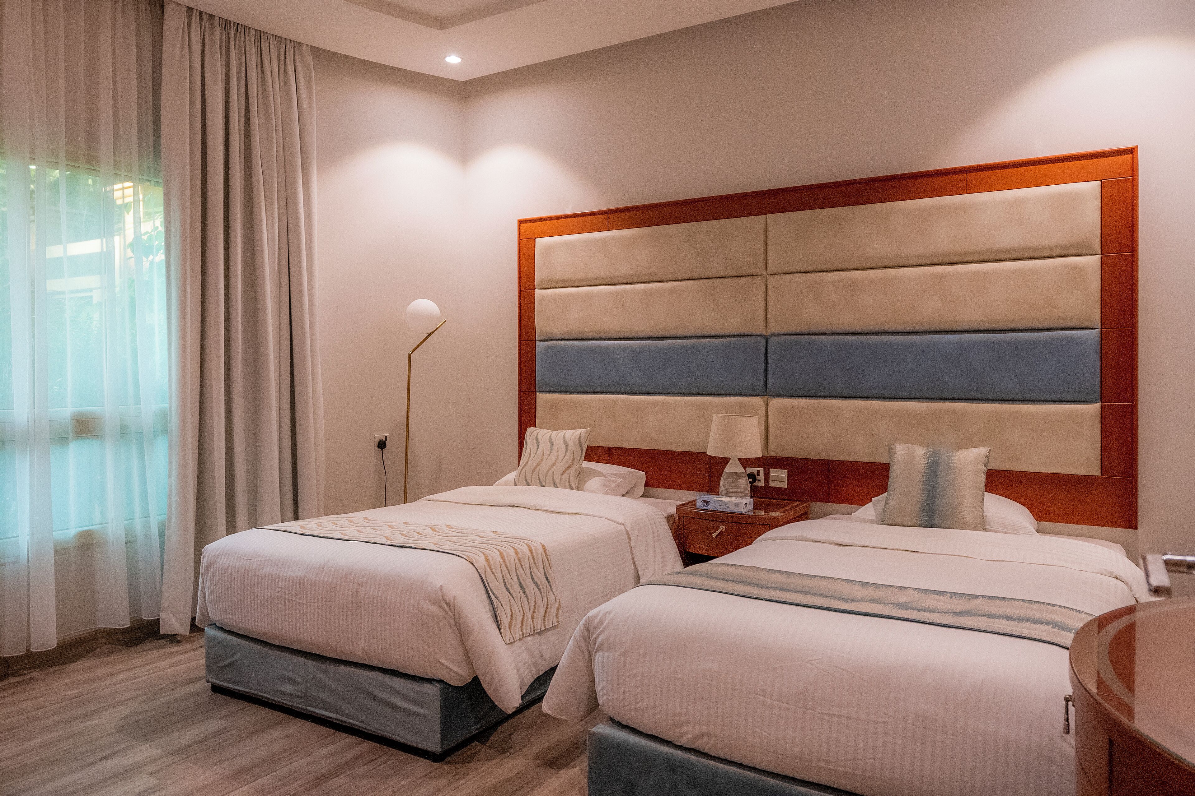 A hotel room with two beds, a wooden headboard, a bedside table with a lamp, and a large window with curtains.