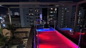 Un espace piscine aménagé sur le toit, doté d’une balustrade en verre, de chaises longues et offrant une vue sur les bâtiments environnants la nuit.