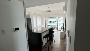 Une cuisine moderne avec un plan de travail noir et des meubles blancs, un coin repas avec deux tabourets de bar, et une grande fenêtre donnant sur un balcon.