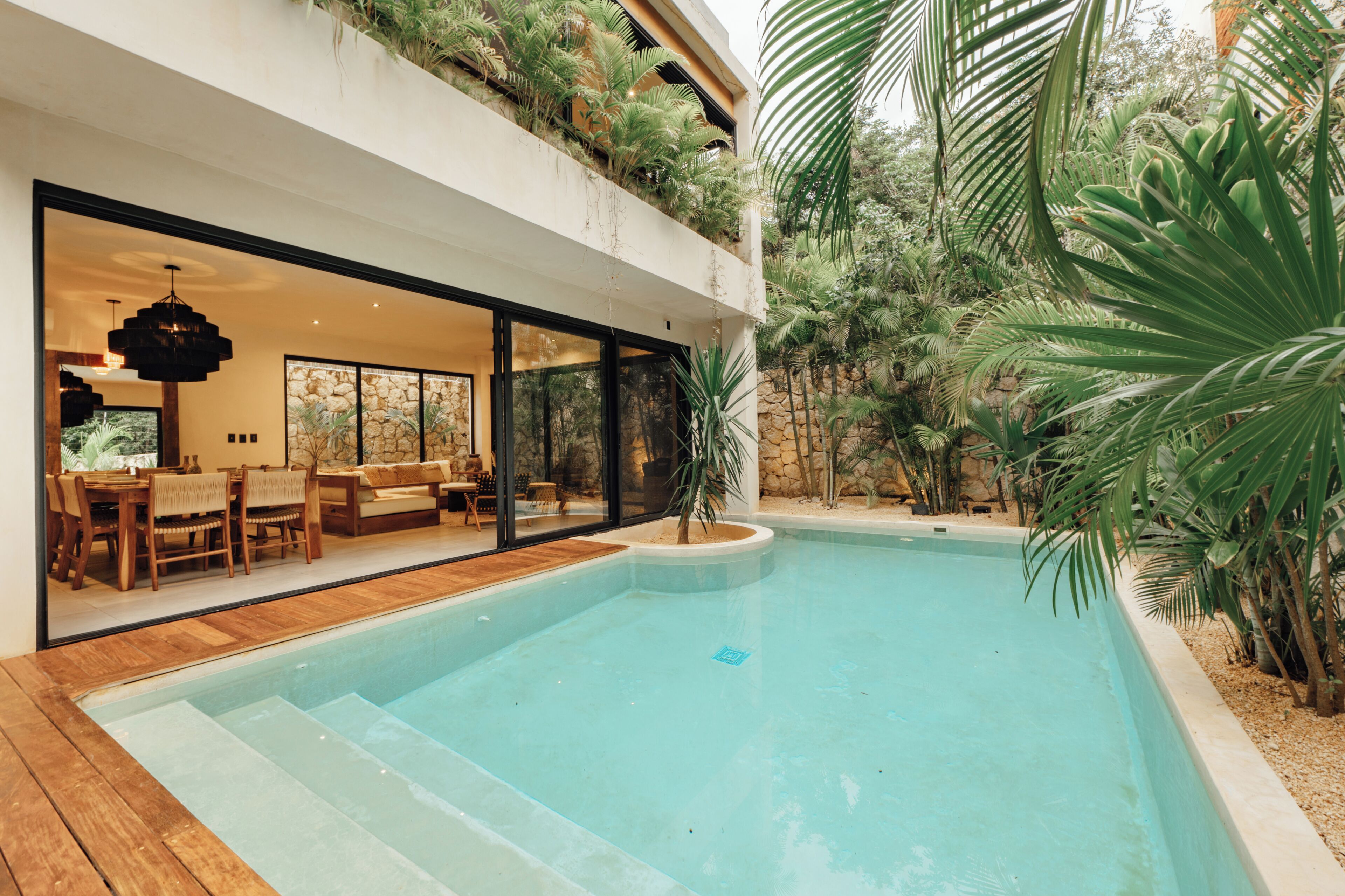Una piscina moderna con terraza de madera, un área de comedor con mesa y sillas, y un amplio ventanal con vistas a la piscina.