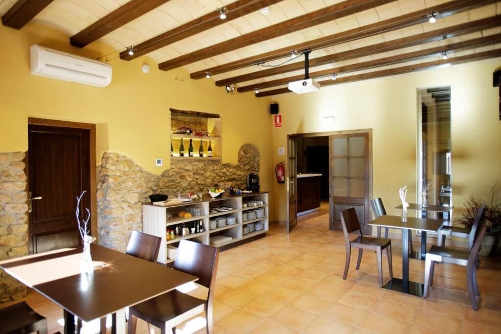 Un espace repas aux murs en pierre, avec des poutres en bois et un sol carrelé. On y trouve des tables et des chaises, ainsi qu’une étagère remplie de bouteilles.