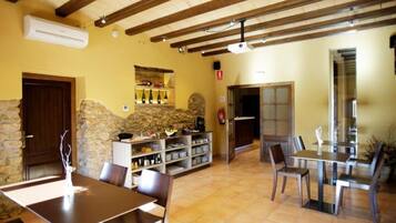 Un espace repas aux murs en pierre, avec des poutres en bois et un sol carrelé. On y trouve des tables et des chaises, ainsi qu’une étagère remplie de bouteilles.