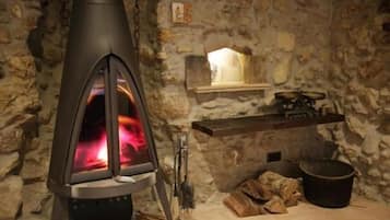 A stone-walled room with a wood-burning stove, a shelf with a candle, and a bucket of firewood.