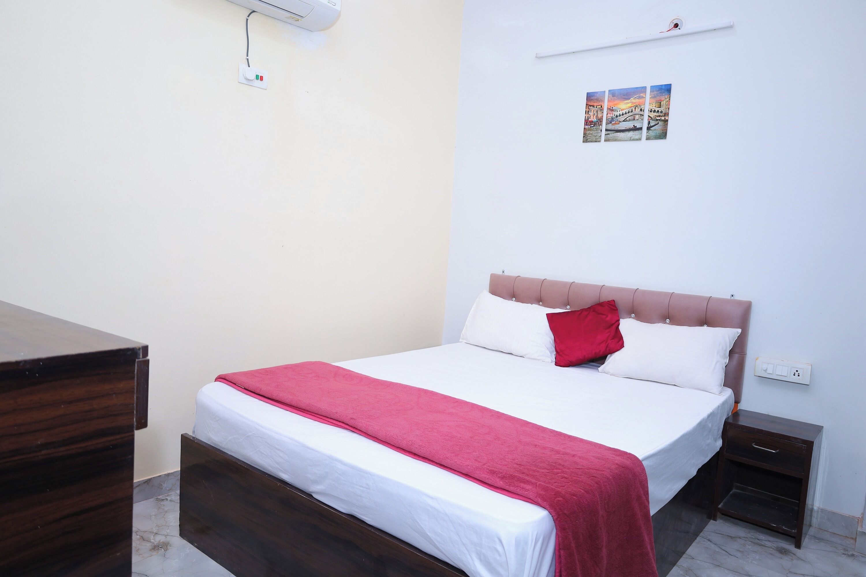 Una cama bien hecha con una manta roja, una mesita de noche de madera y un estante en la pared con fotografías.