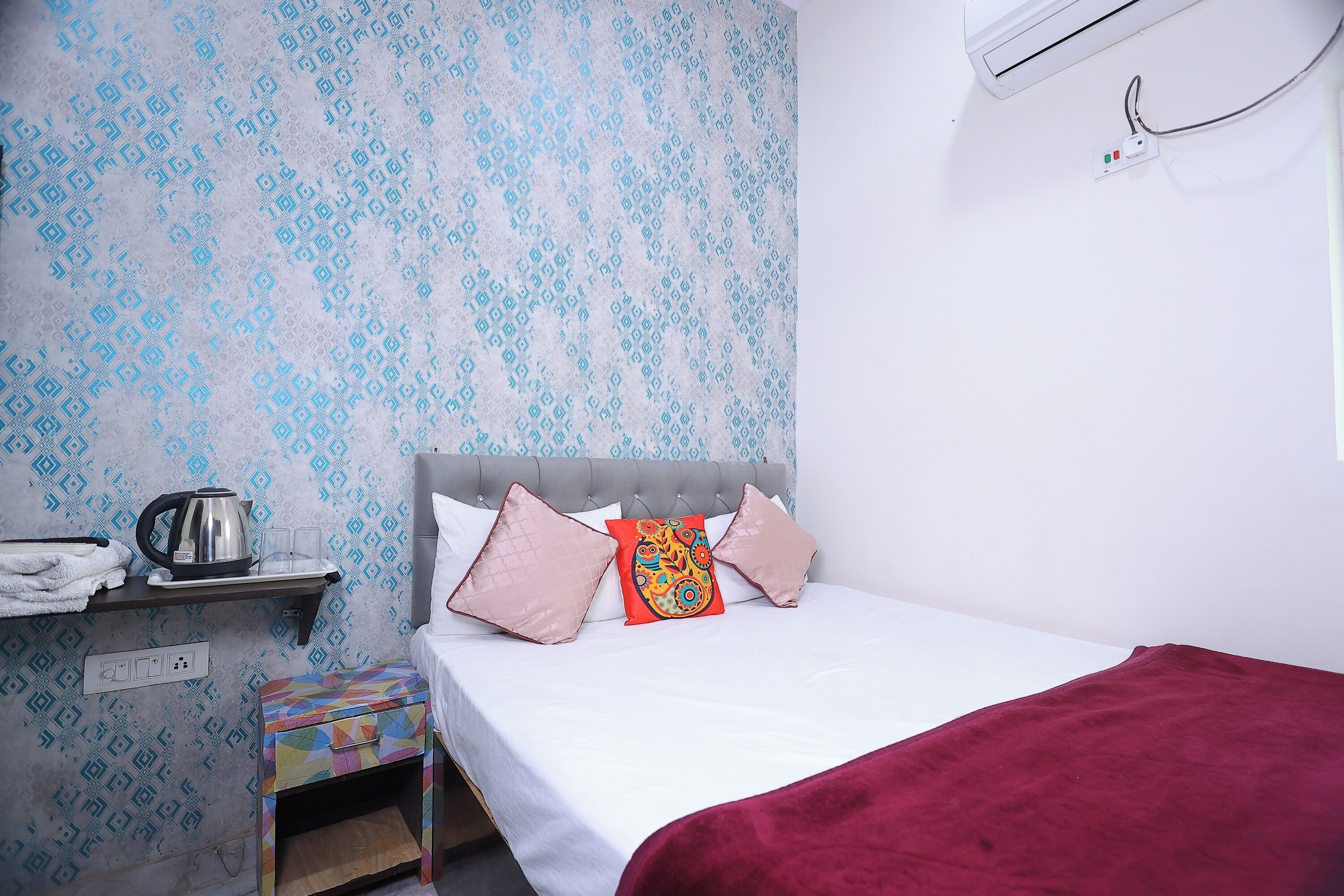 A bedroom with a bed, a nightstand, a kettle, and a power outlet.
