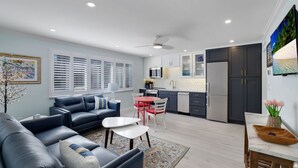 Una sala de estar moderna con un sofá azul, una mesita de centro pequeña y un comedor con una mesa roja y sillas blancas. La cocina cuenta con armarios oscuros, un refrigerador y un salpicadero de azulejos.