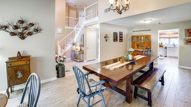 Une salle à manger spacieuse, avec une grande table en bois, des chaises bleues et un lustre. Un escalier mène à l’étage supérieur.