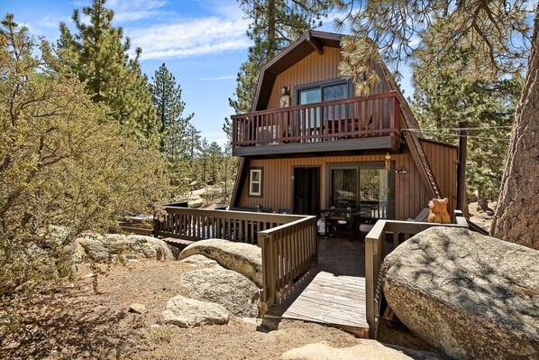 A two-story cabin with a balcony, surrounded by trees and rocks.