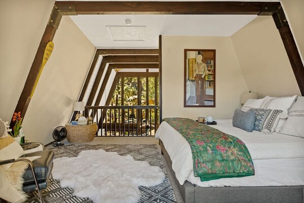 Un dormitorio con techo estilo loft, una cama con colcha floral, una alfombra de piel blanca, una mesita con un ventilador y vistas al exterior desde un balcón.